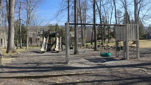 A playground with swings and a slide, surrounded by trees and a building in the background. - Pocono Mountains Escape - 2 Bedroom, 2.5 Bath, private townhome with wood-burning fireplace (East Stroudsburg)