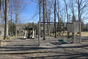 Un parque infantil con columpios y un tobogán, rodeado de árboles y un edificio al fondo.