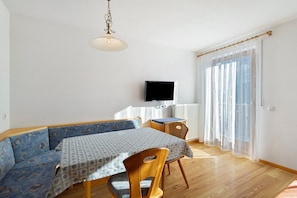 A room with a wooden bench, a table with a patterned tablecloth, a television on the wall, and sheer curtains on the window.