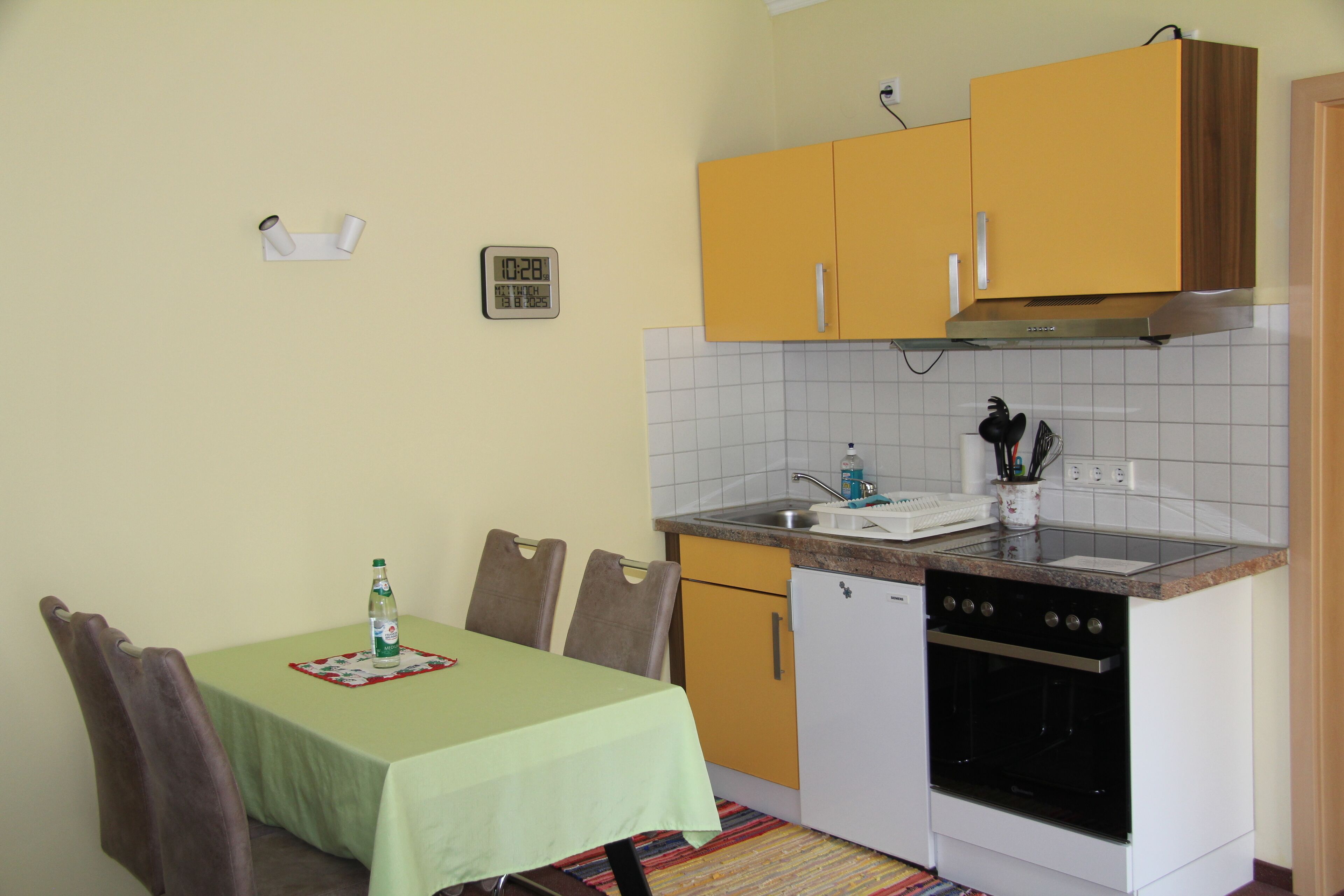 A small kitchen with a dining table, chairs, a refrigerator, an oven, and a clock on the wall.