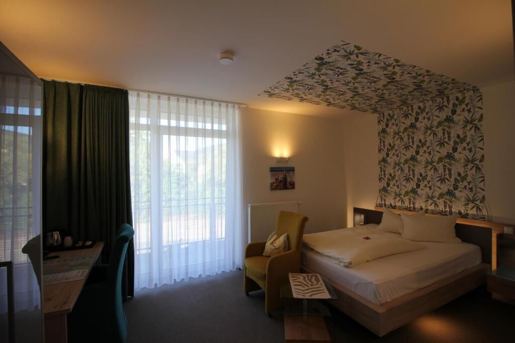 A hotel room with a bed, a chair, a desk, and a large window with sheer curtains.