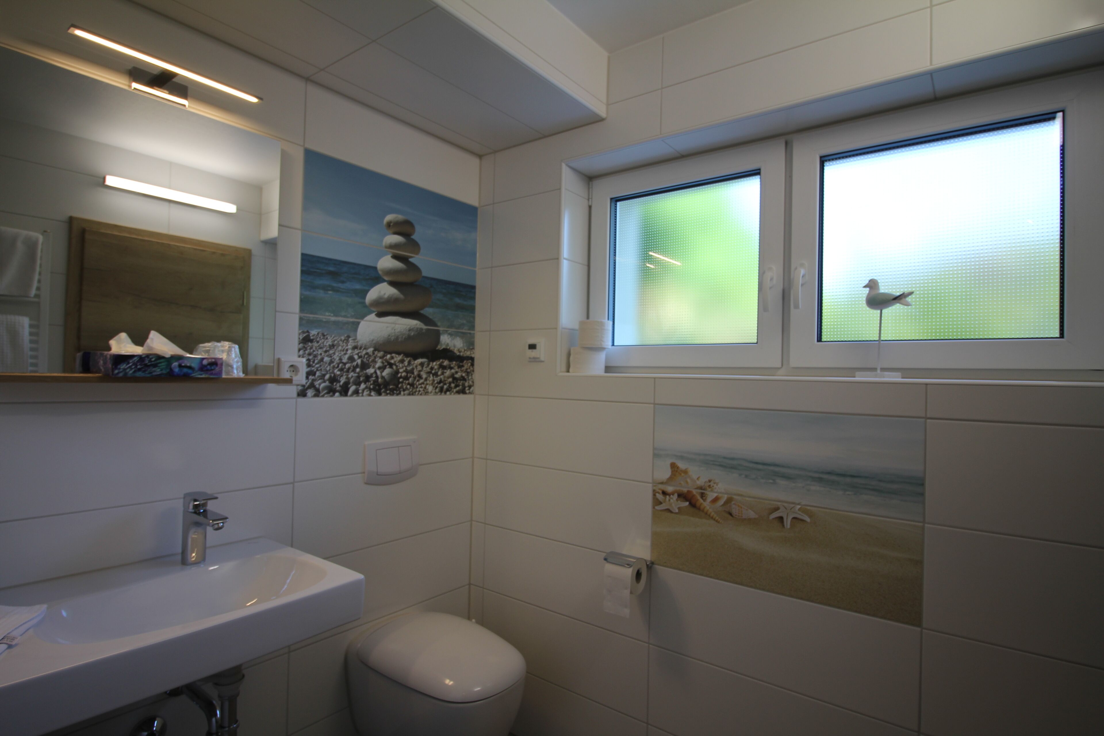 Een badkamer met een wastafel, toilet en decoratieve tegels, waaronder een afbeelding van een strand en een vogel.