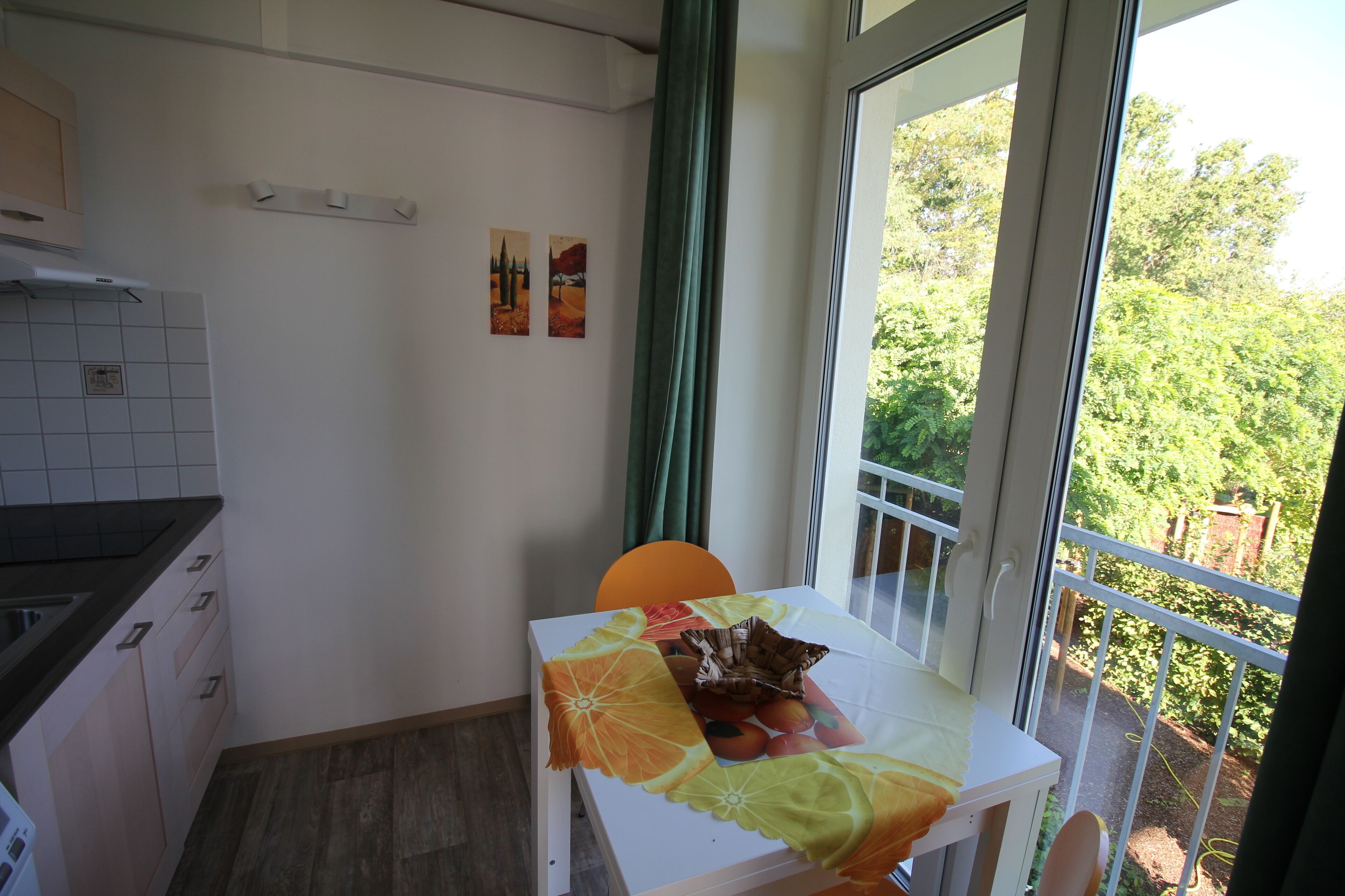 A small dining area with a table set for two, a window with a view of greenery, and a kitchen area with cabinets and a countertop.