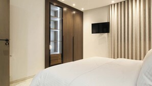 A bedroom with a large bed, a wardrobe with shelves, a flat-screen TV mounted on the wall, and a window with vertical blinds. - Al Yassmine 5-6 Apartment - A True Gem (Sfax)