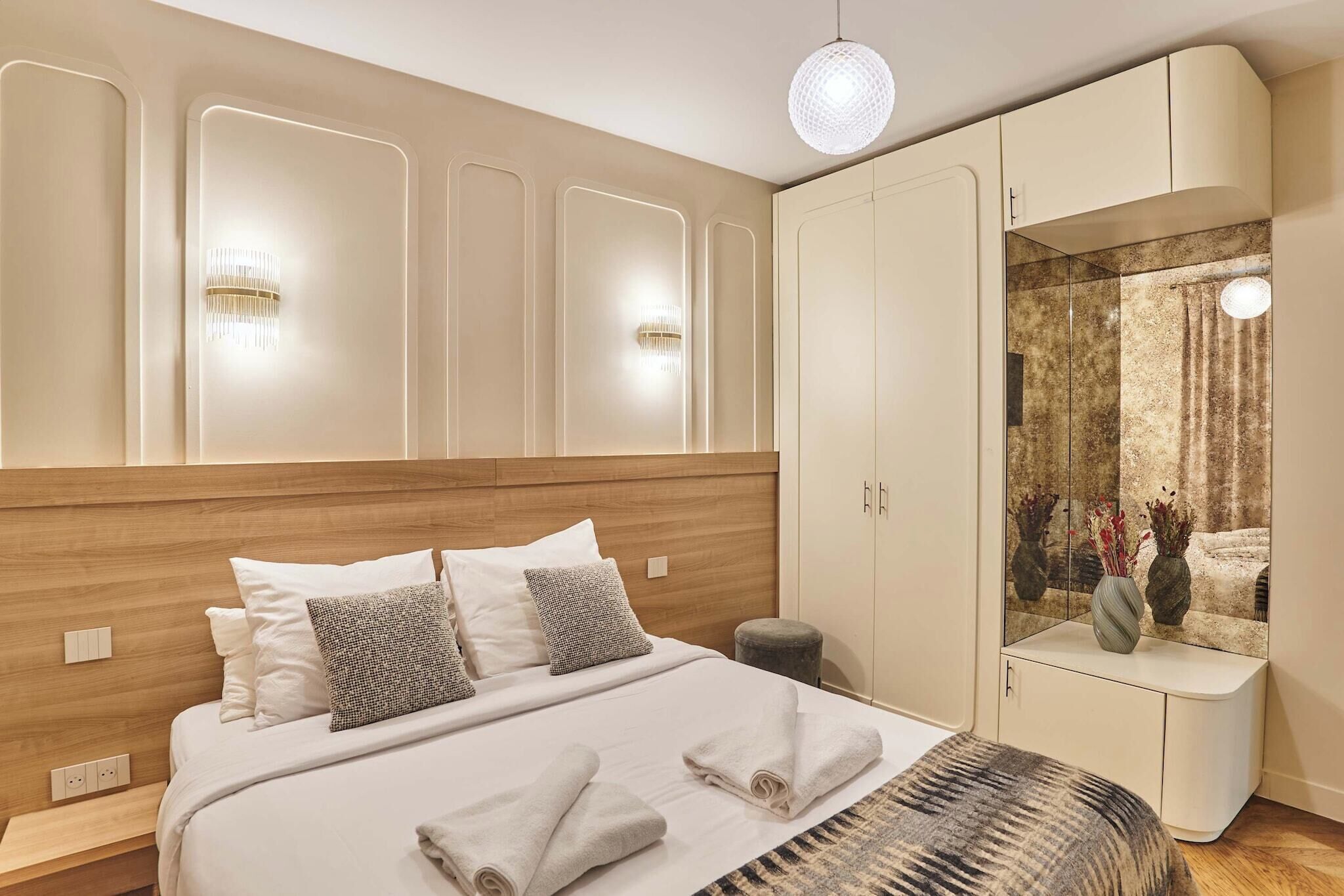 Ein modernes Schlafzimmer mit einem hölzernen Kopfteil, einem großen Bett mit weißer Bettwäsche, einem Nachttisch und einem Kleiderschrank mit Spiegel.