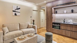 A modern living room with a beige sofa, a coffee table, and a kitchen area with wooden cabinets and a built-in oven. - Superb air-conditioned flat on the Champs Elysées (Paris)