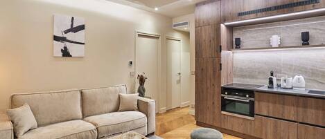 A modern living room with a beige sofa, a coffee table, and a kitchen area with wooden cabinets and a built-in oven.
