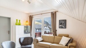 Ein Wohnzimmer mit einer Couch, zwei Sesseln, einem Couchtisch, einer Tür, einem Fenster mit Vorhängen und einem gerahmten Bild an der Wand.