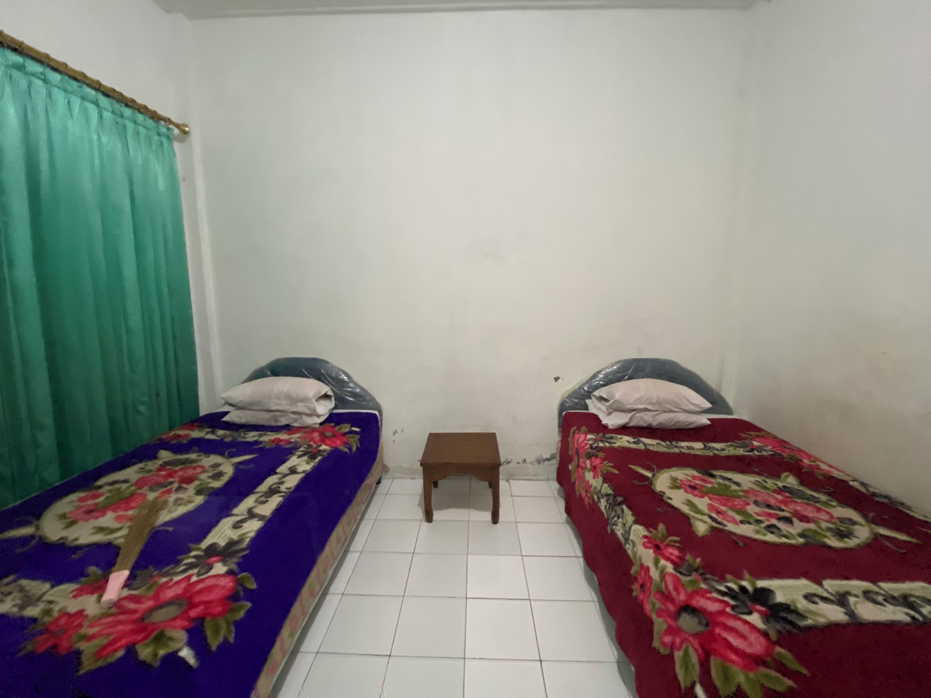A small, clean room with two single beds, a small wooden table, and a window with curtains.