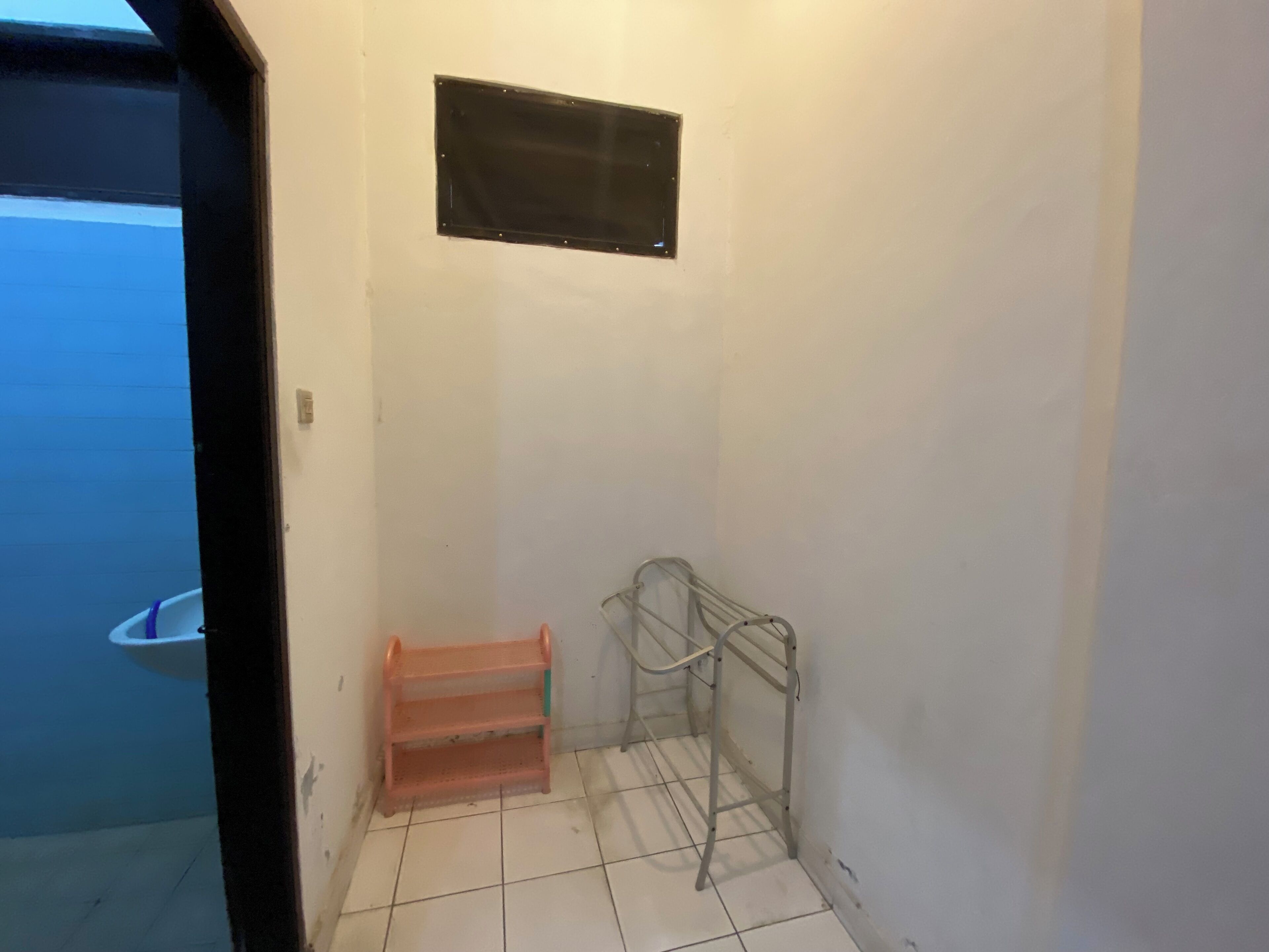 A small, enclosed bathroom with a tiled floor, a metal shelf, and a small window.