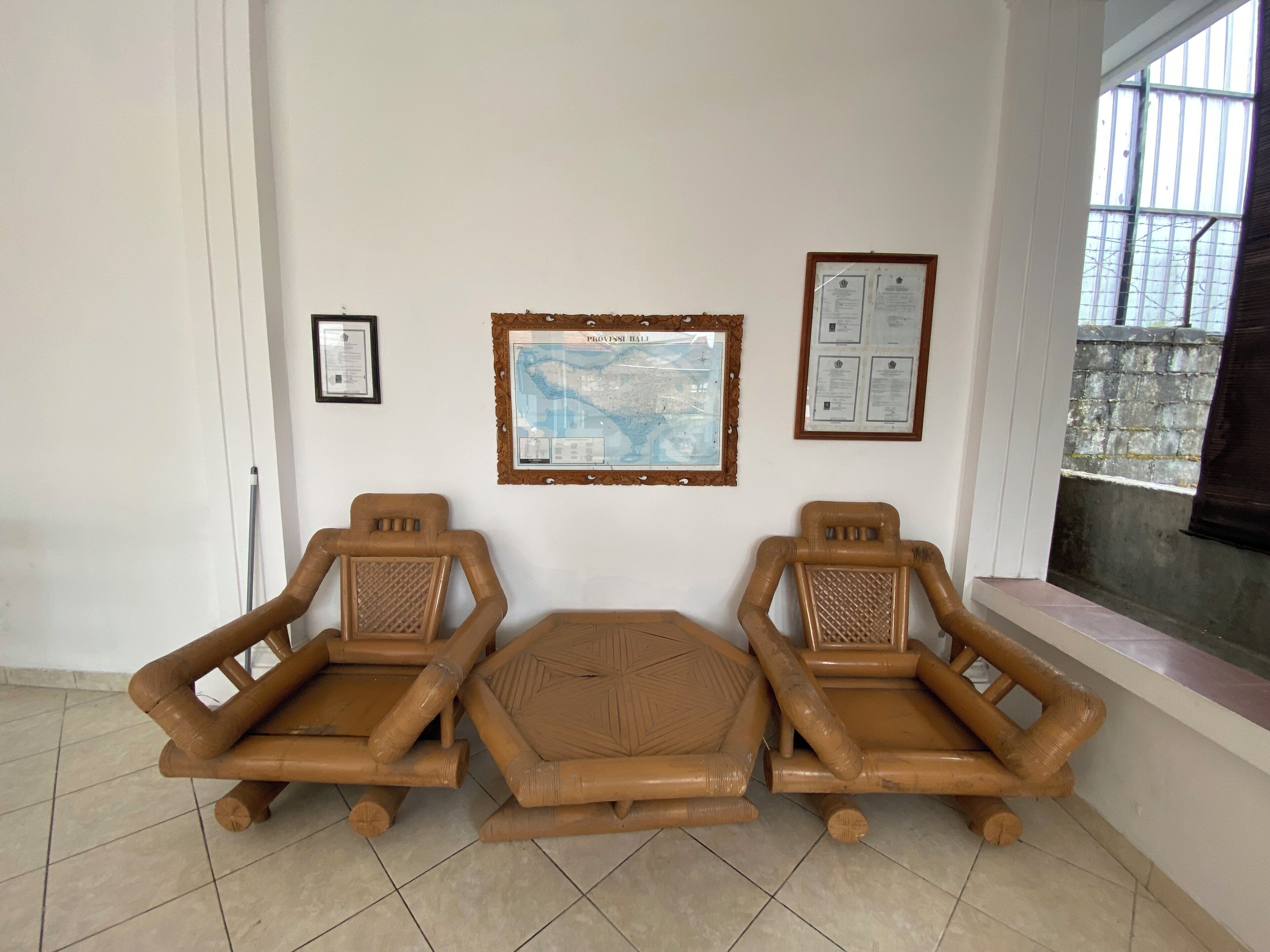 A room with three bamboo chairs, a framed map on the wall, and a window with a view of a stone wall.