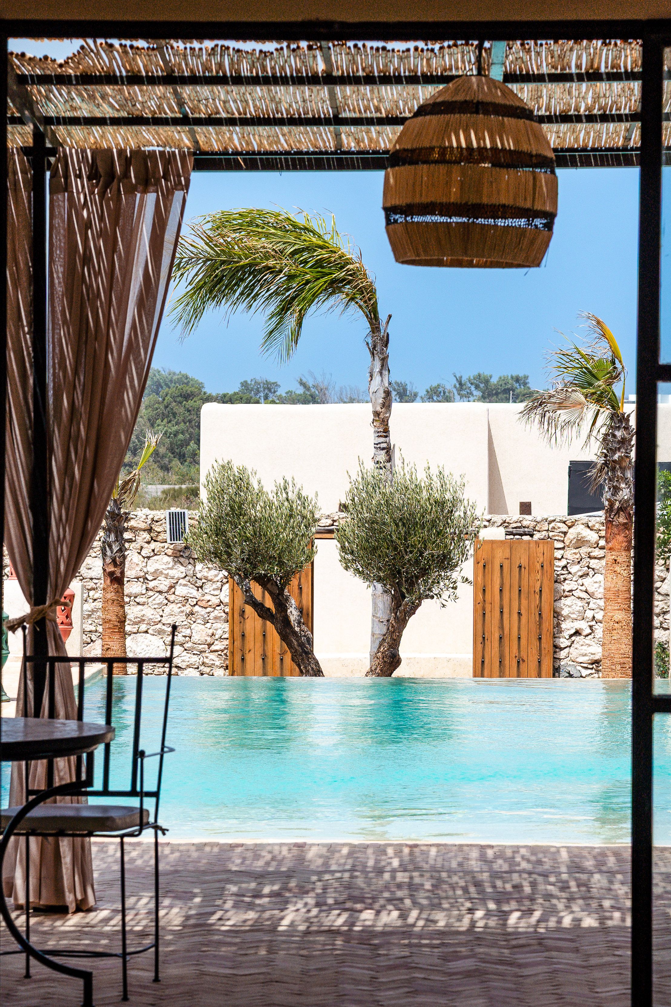 Un área de piscina con un pérgola, palmeras y un muro de piedra.