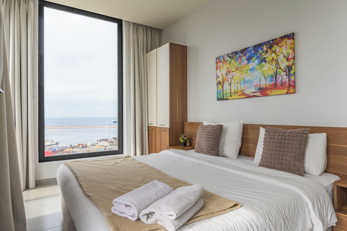 A hotel room with a large window, a bed with white linens, a wooden headboard, and a painting of trees.
