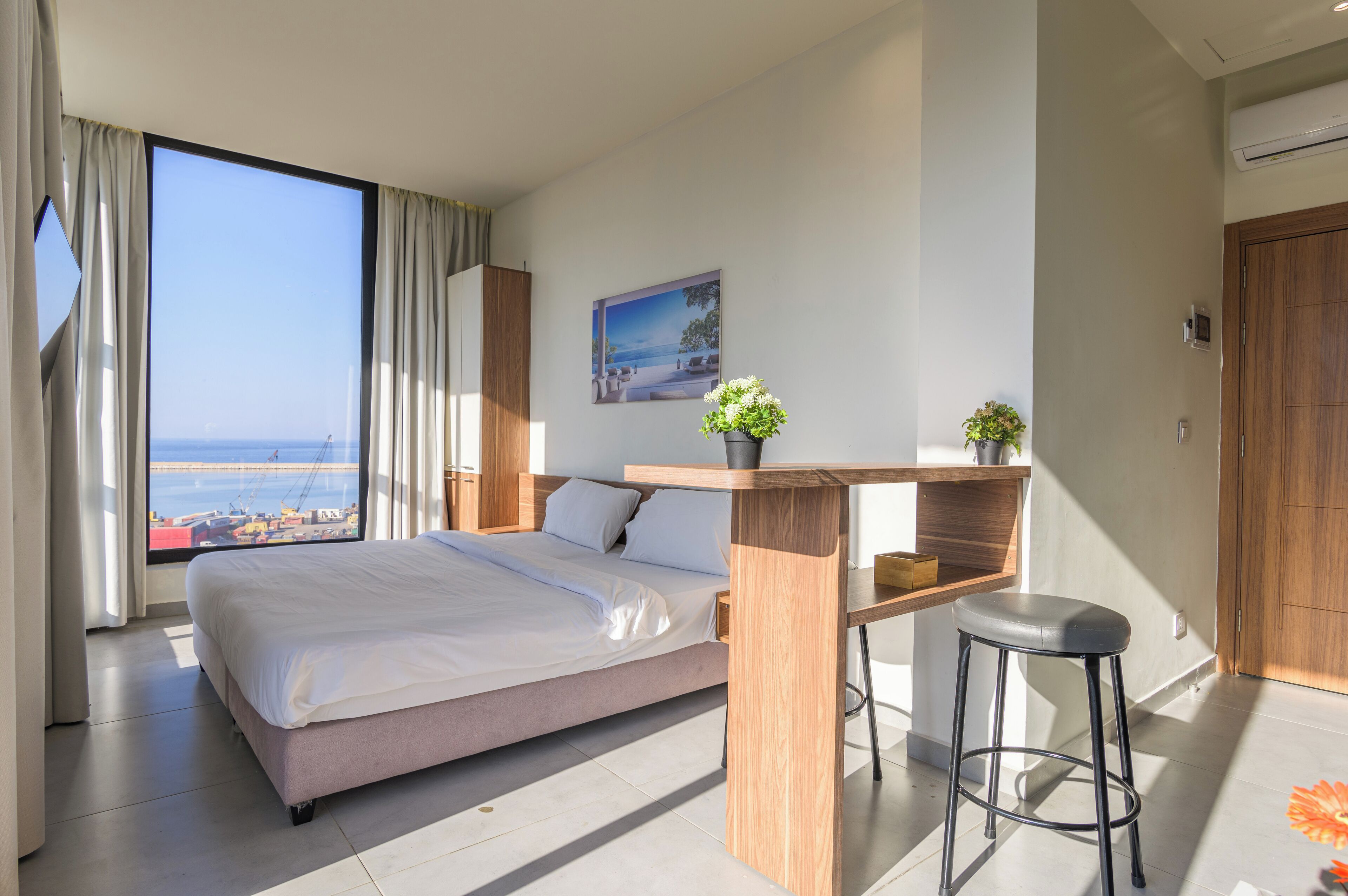 A modern hotel room with a large bed, a desk with a chair, a view of the sea, and a potted plant.