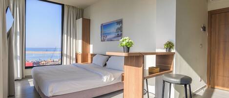 A modern hotel room with a large bed, a desk with a chair, a view of the sea, and a potted plant.