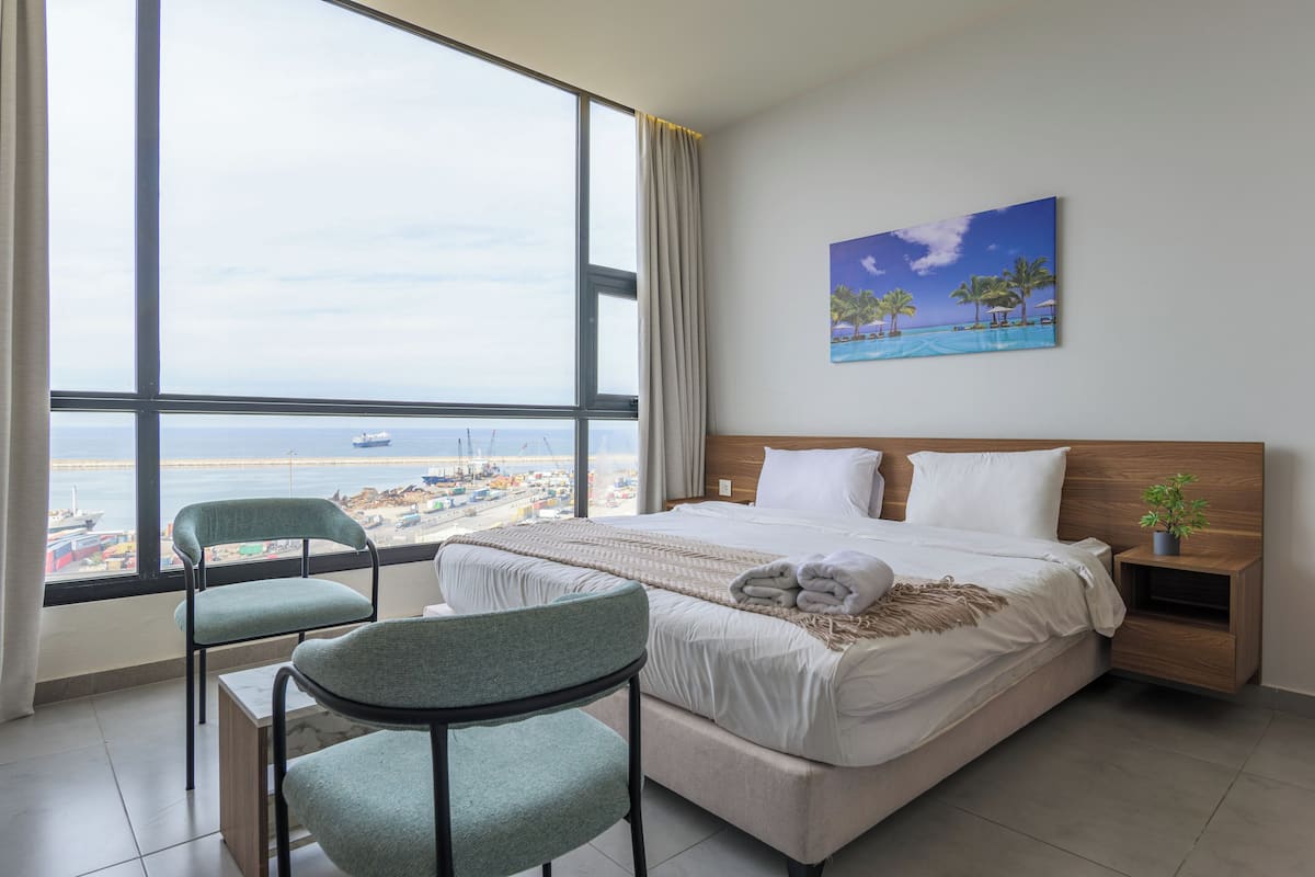 A hotel room with a large window, a bed, two chairs, a nightstand, and a painting of a beach scene.