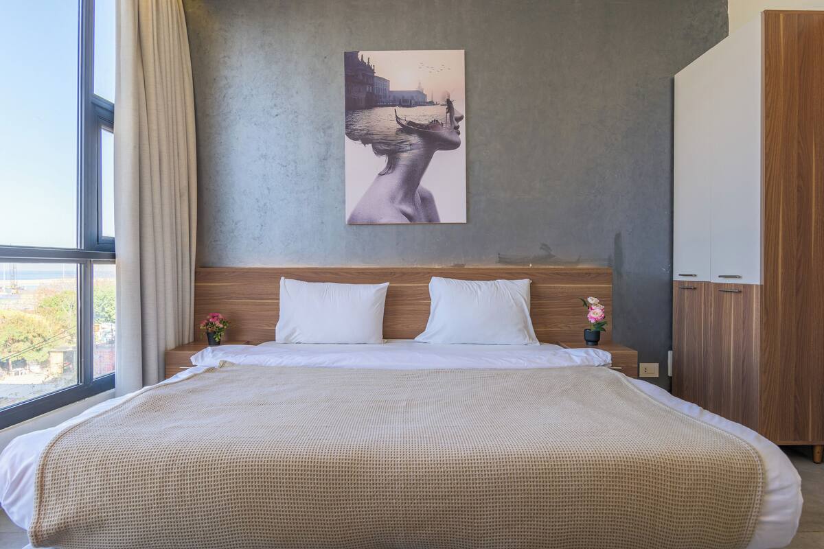 A modern hotel room with a large bed, a wooden headboard, a painting of a cityscape, and a view of the outdoors.