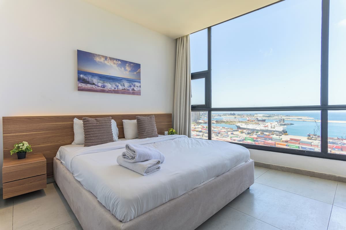A modern hotel room with a large bed, a nightstand, a potted plant, and a painting of a beach scene.