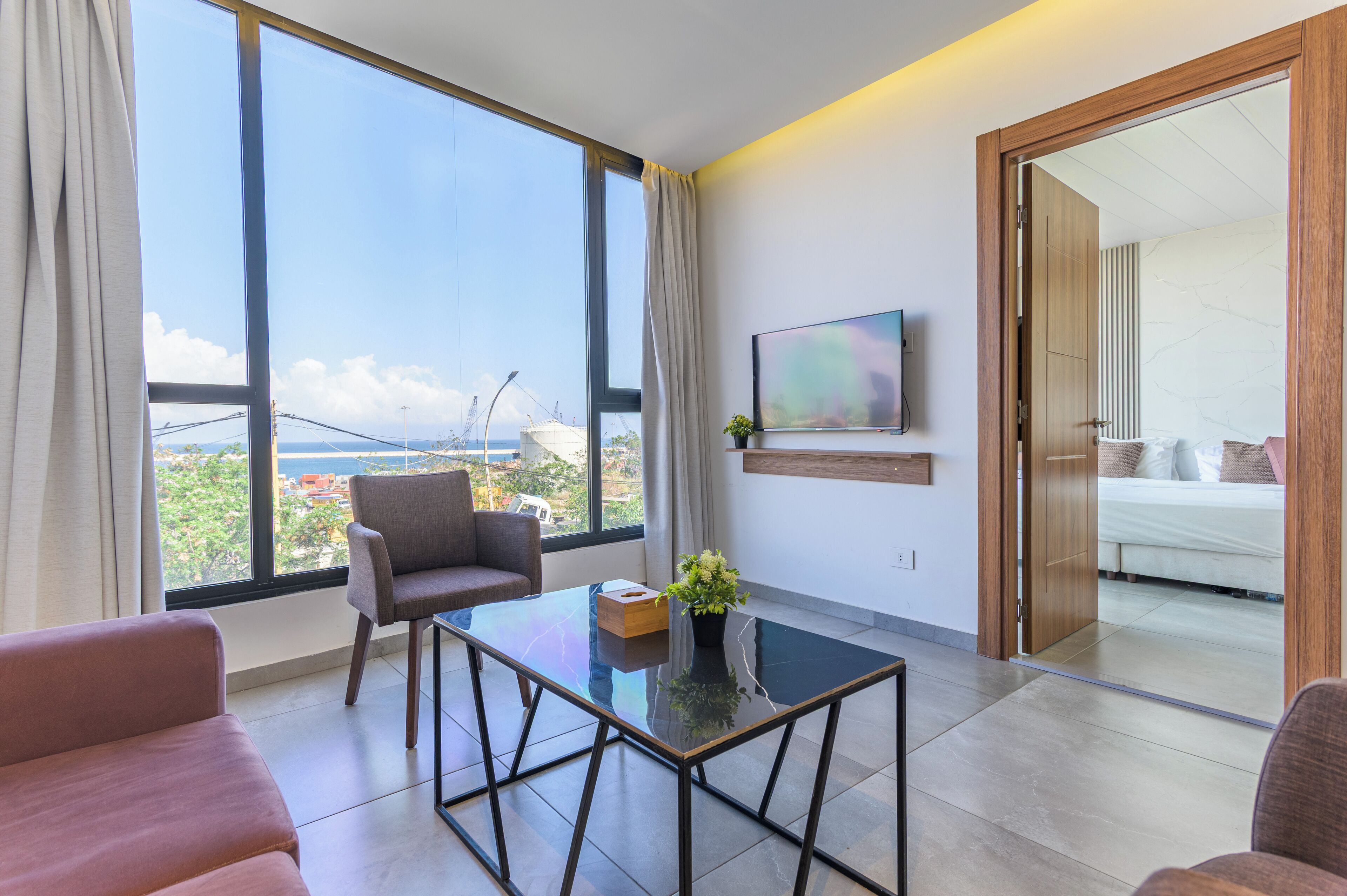 Una habitaci ón de hotel moderna con un ventanal grande, una mesa de centro de vidrio, un sofá y una cama con vistas a la ciudad.