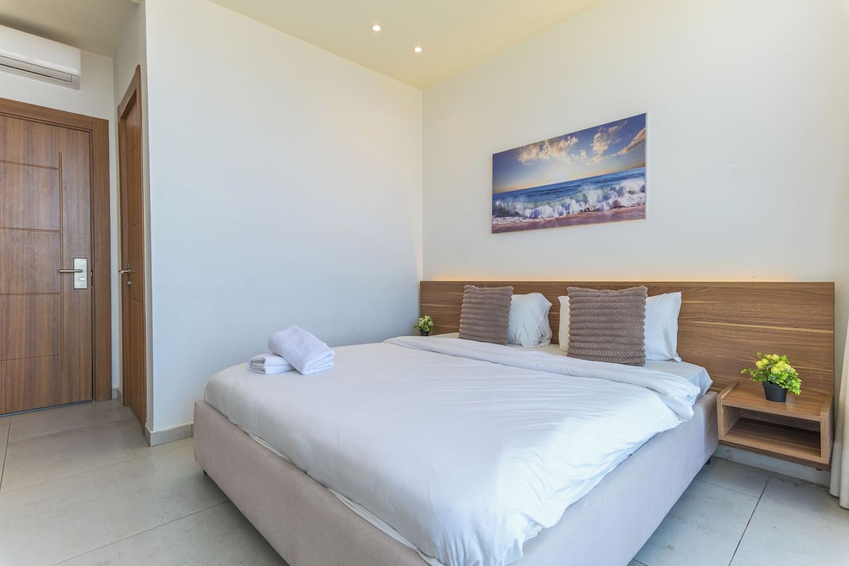 A modern bedroom with a large bed, a wooden headboard, a nightstand with a plant, and a painting of a beach scene on the wall.