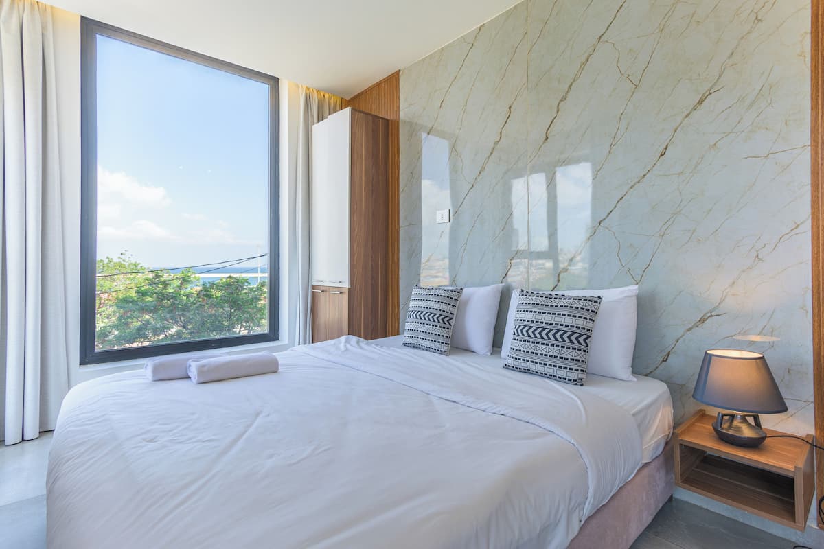 A hotel room with a large bed, a nightstand, a lamp, and a view of the outdoors.