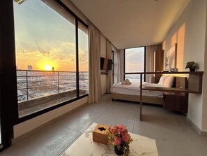 Ein Hotelzimmer mit Bett, Schreibtisch, großem Fenster mit Stadtblick, Pflanze und Uhr.