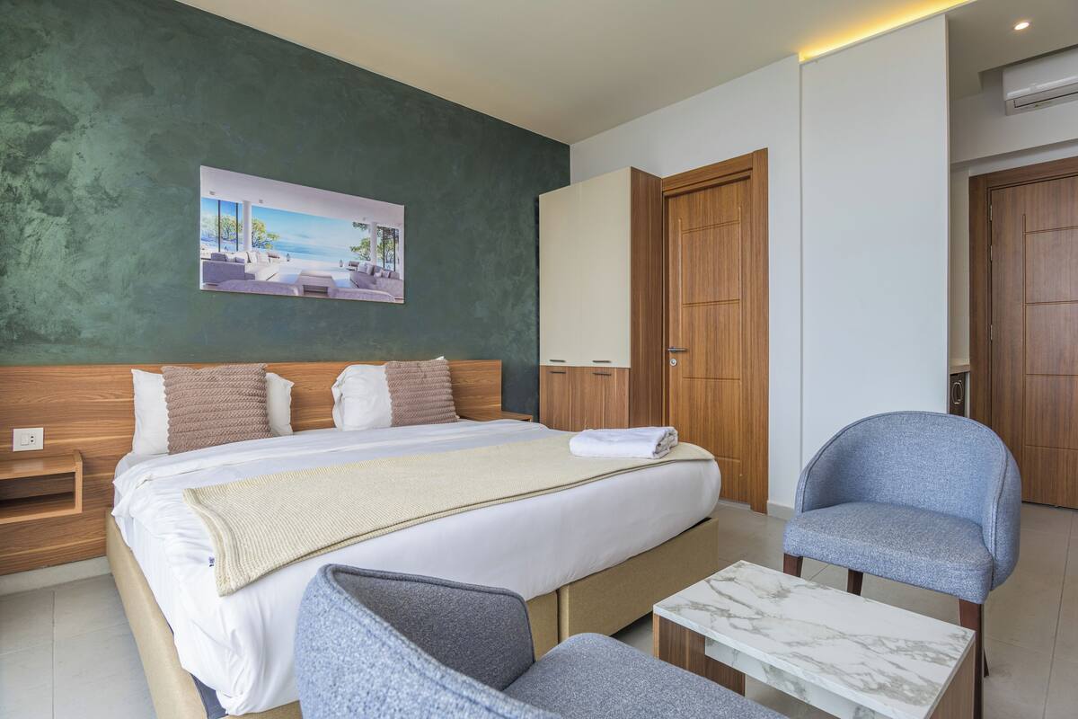 A modern hotel room with a large bed, a wooden wardrobe, a seating area with a chair and a small table, and a painting on the wall.