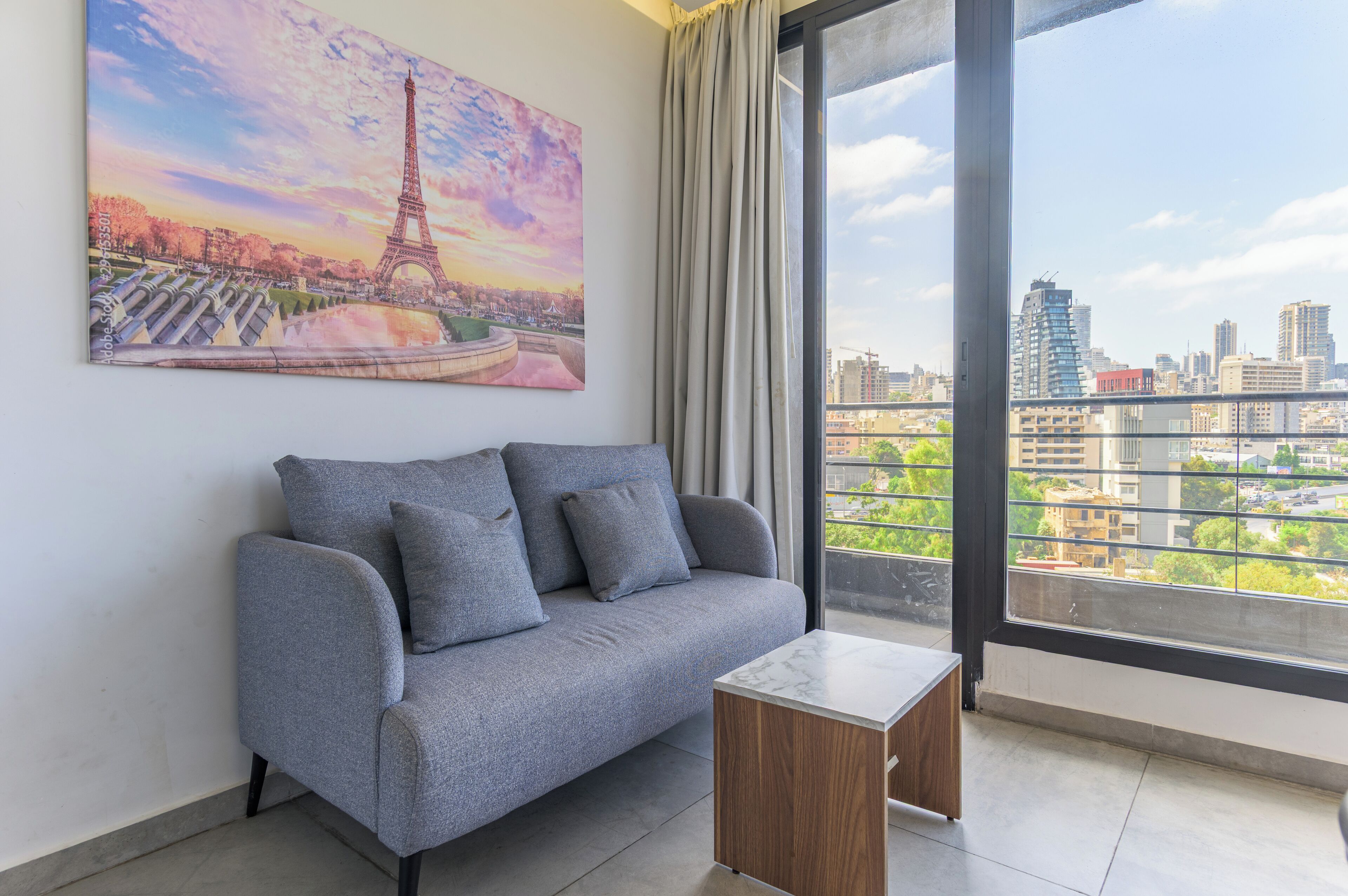 A modern living room with a grey sofa, a small wooden side table, and a large window offering a cityscape view.