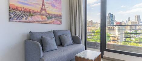 A modern living room with a grey sofa, a small wooden side table, and a large window offering a cityscape view.