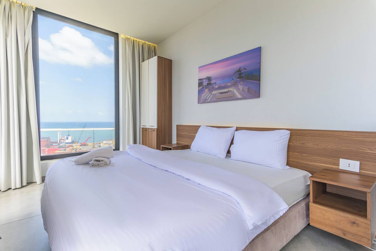 A hotel room with a large bed, a wooden headboard, a painting of a sunset, and a view of the sea through a window.