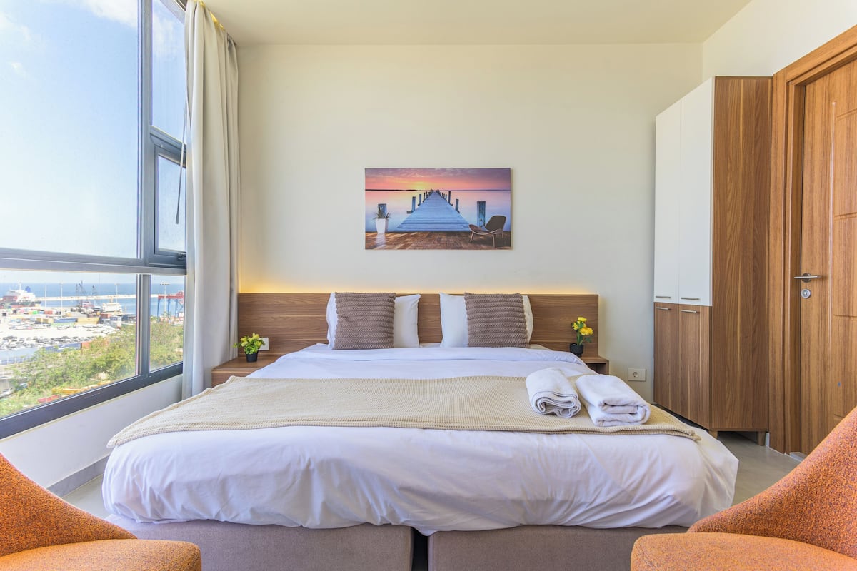 A hotel room with a large bed, a nightstand, a wooden wardrobe, and a window with a view of boats and greenery.