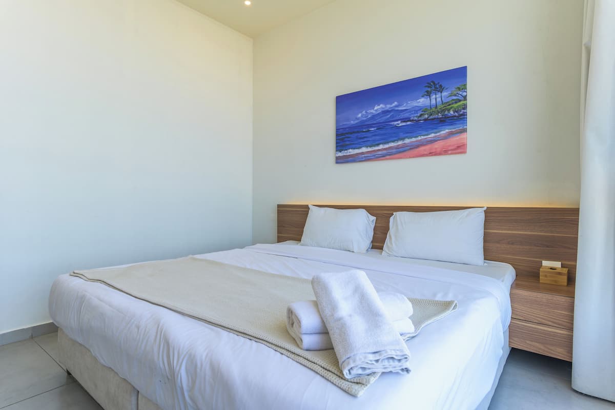 A neatly made bed with white linens and a beige throw blanket, a wooden headboard, and a painting of a beach scene on the wall.