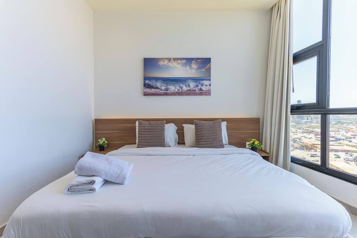 A hotel room with a large bed, a painting of a beach scene, a city view through the window, and a small plant on the nightstand.