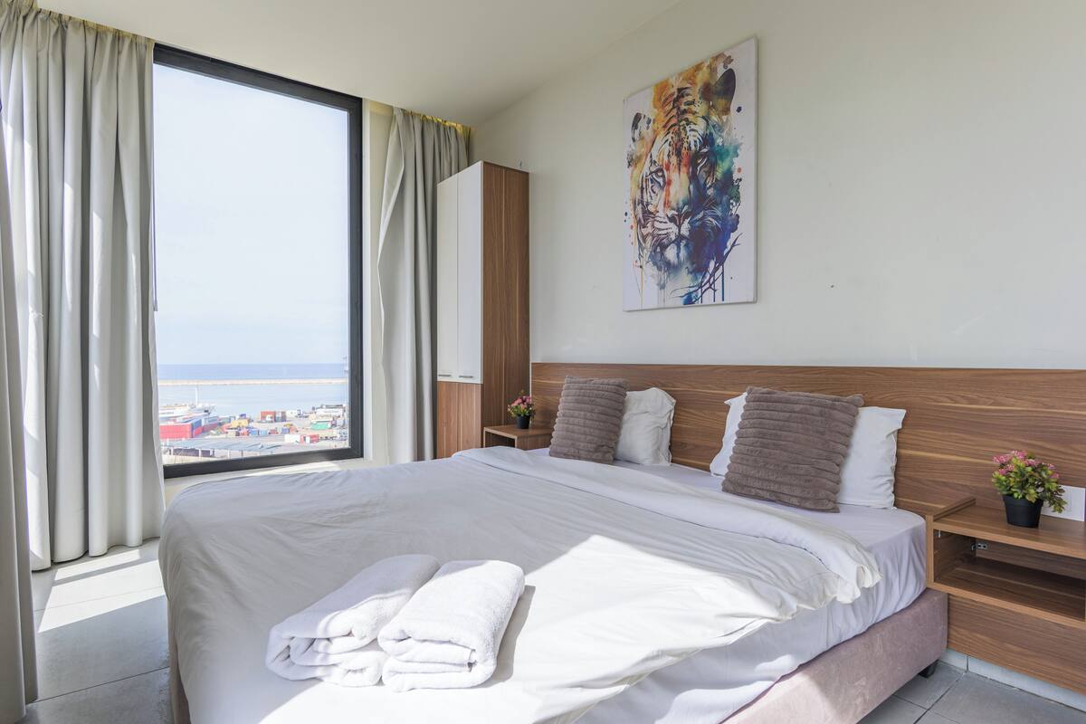 A hotel room with a large bed, a wooden headboard, a painting on the wall, and a view of the sea through the window.
