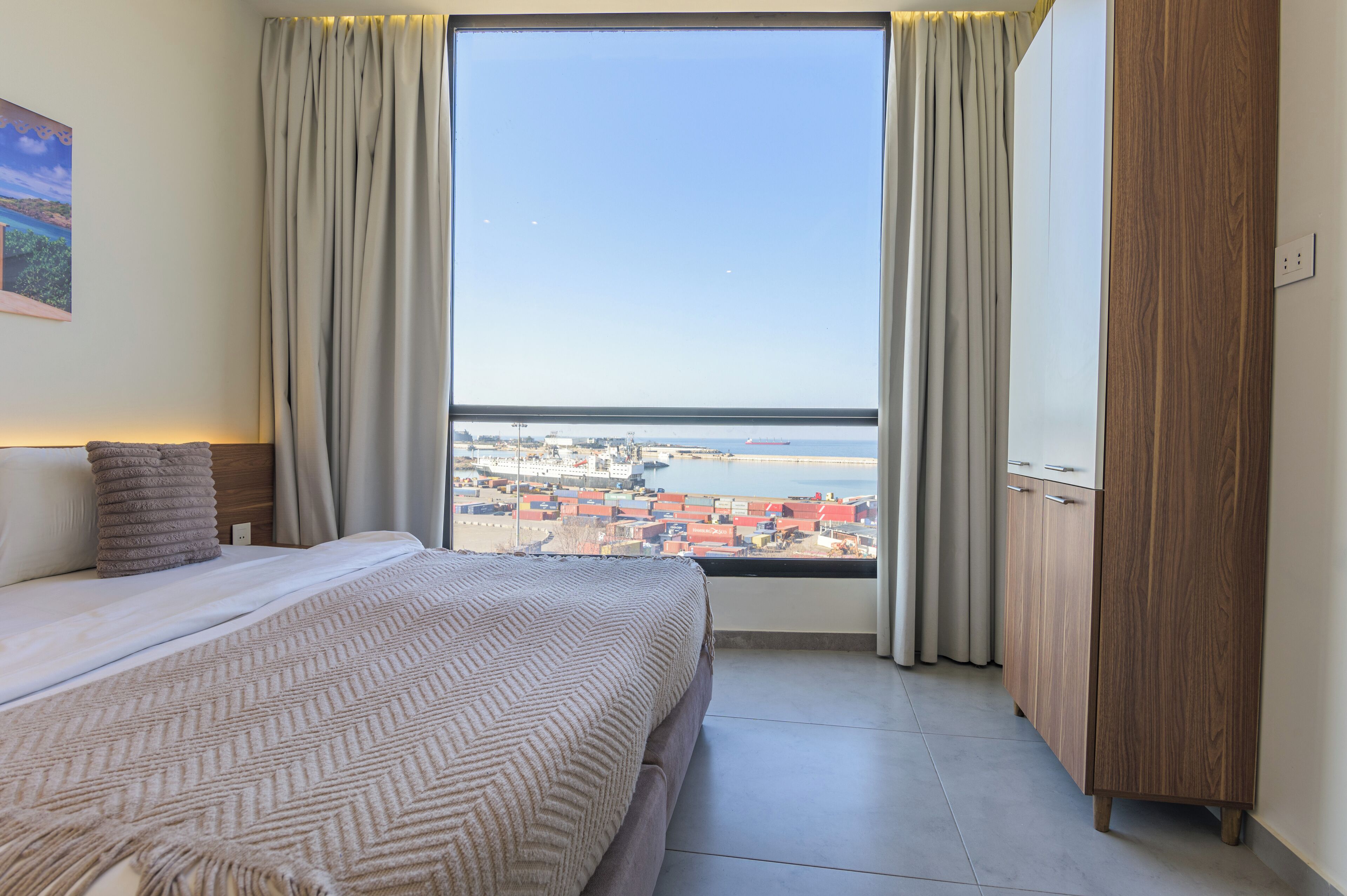 A bedroom with a large window, a bed, a wardrobe, and a view of a cityscape.