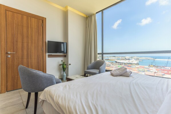 A hotel room with a large bed, a TV, a chair, and a view of the sea and cityscape. - Gate 9 Tower -Studios & Apts Mar Mikhael (Beirut)