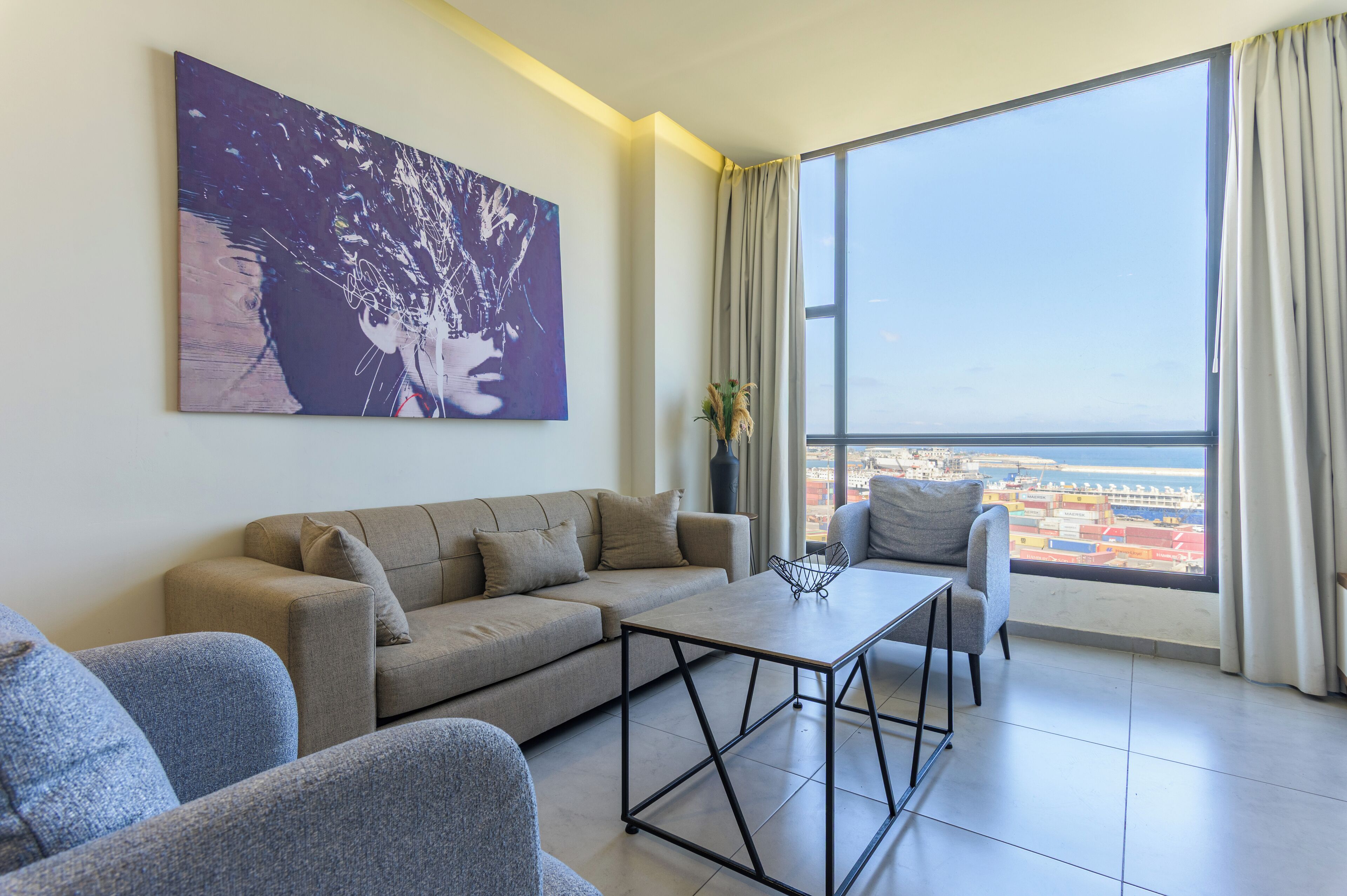 A modern living room with a sofa, armchairs, a coffee table, and a large window offering a city view.