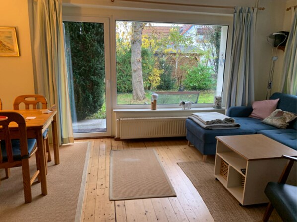 A living room with a wooden floor, a blue sofa, a small table, and a window with a view of trees and a garden.