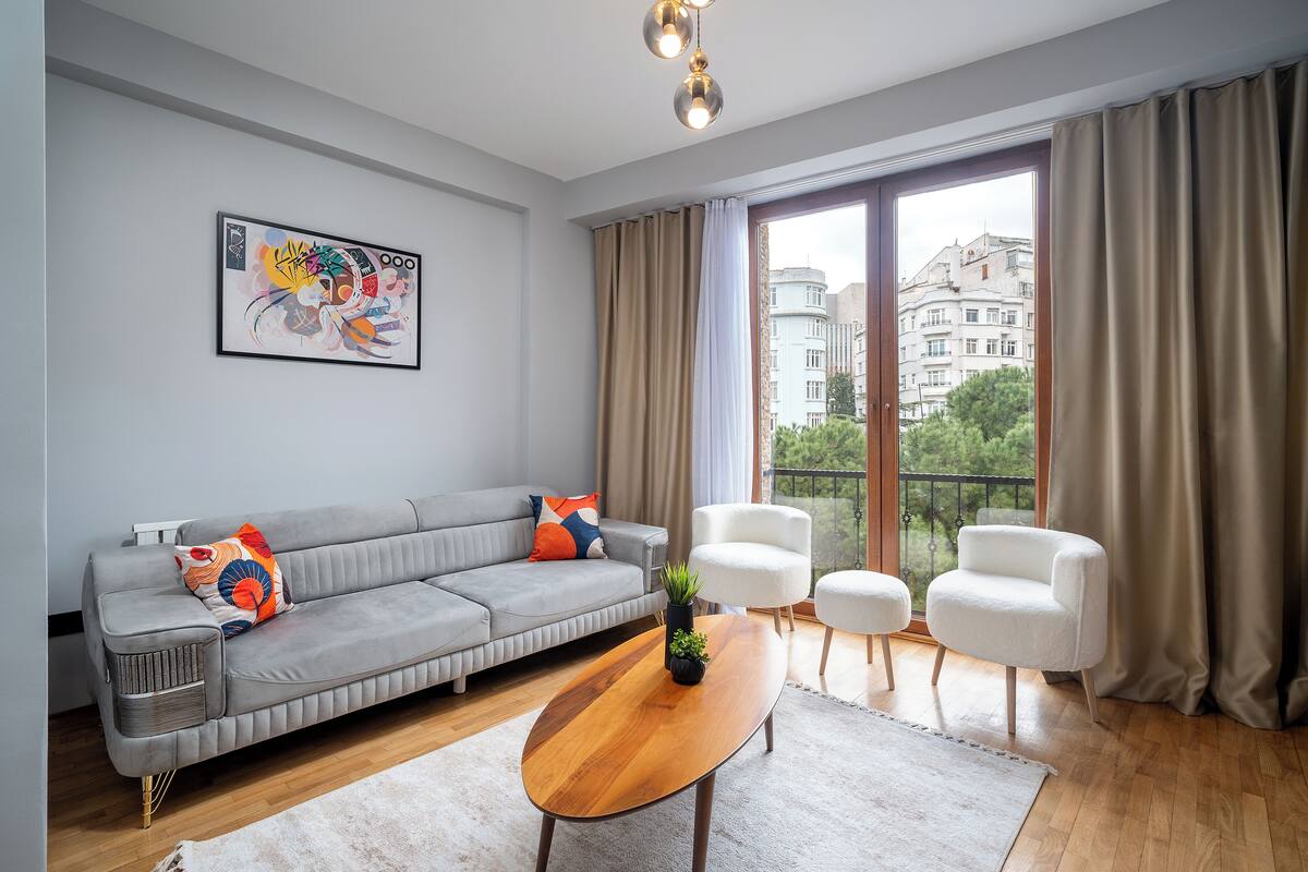 Una sala de estar moderna con un sofá gris, sillones blancos, una mesa de centro de madera y un ventanal con cortinas.