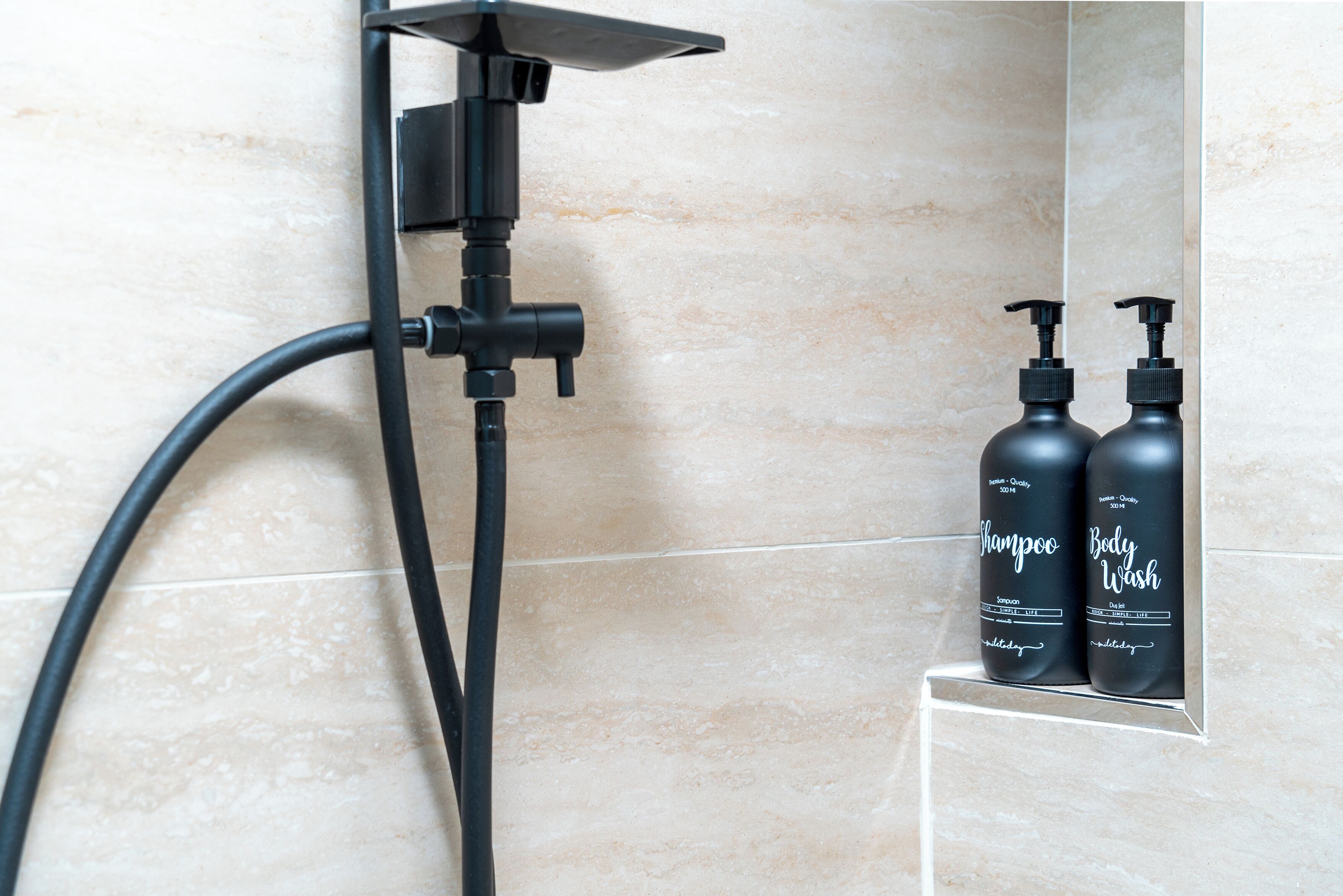 A bathroom with a shower, a handheld showerhead, and two bottles of shampoo and body wash on a shelf.