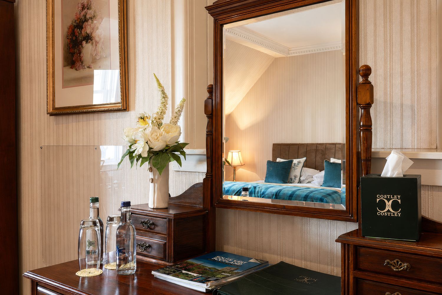 Habitación de hotel con un espejo grande, una mesita de noche con un jarrón de flores, una lámpara y una cama con ropa de cama azul.