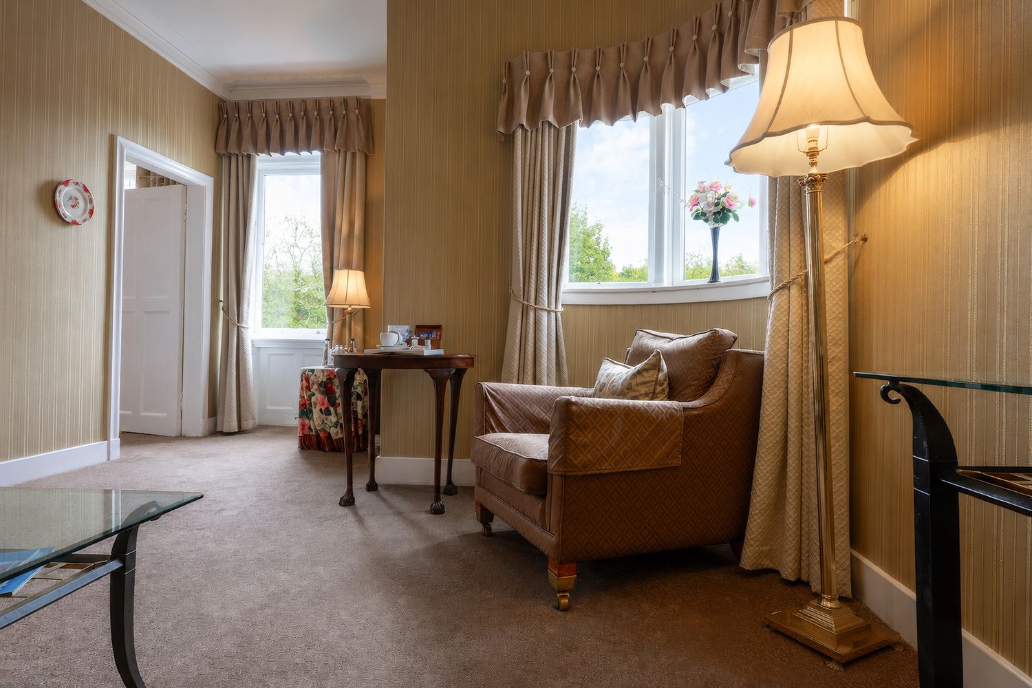 A room with a brown leather sofa, a small table with a lamp, and a window with curtains.