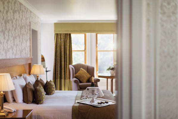 A hotel room with a neatly made bed, a side table with a lamp, a chair, and a small table with a bottle and glasses. - Lochgreen House Hotel & Spa (Troon)