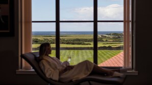 Une personne assise près d’une fenêtre, avec vue sur un paysage verdoyant et un bâtiment situé au loin.