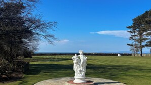Une statue se dresse sur une plateforme circulaire en pierre, entourée d’une pelouse bien entretenue. Un ciel bleu clair et des montagnes lointaines se dessinent à l’arrière-plan.