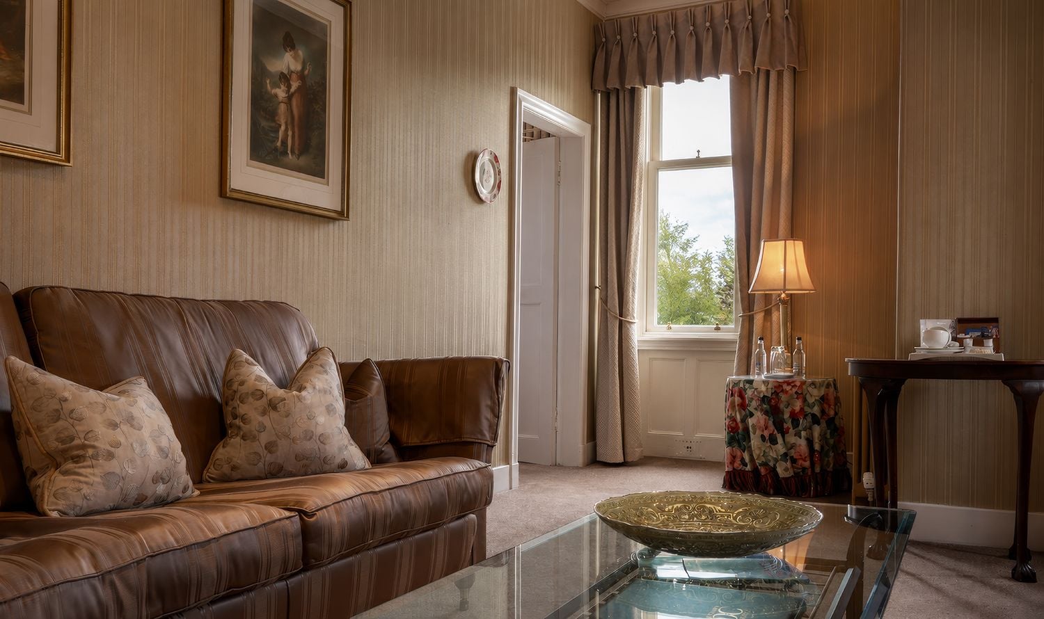 A living room with a brown leather sofa, a glass coffee table, a framed picture on the wall, a window with curtains, and a lamp on a side table.