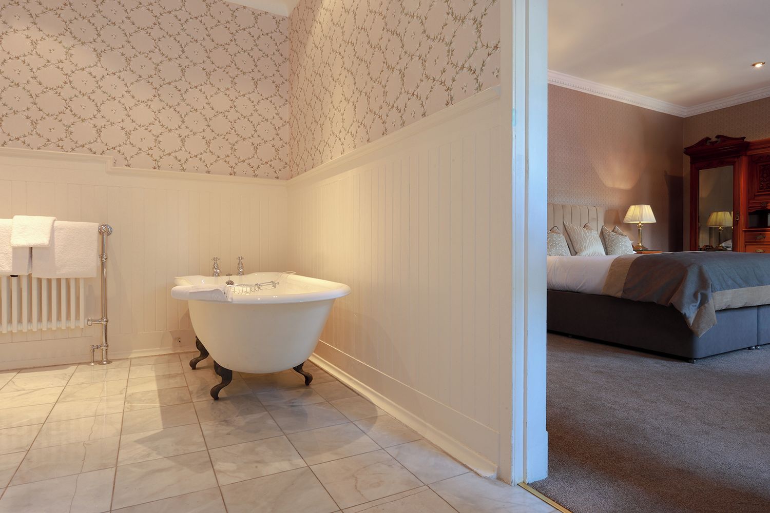 A bathroom with a freestanding bathtub, a radiator, and a bed with a nightstand in the adjacent room.
