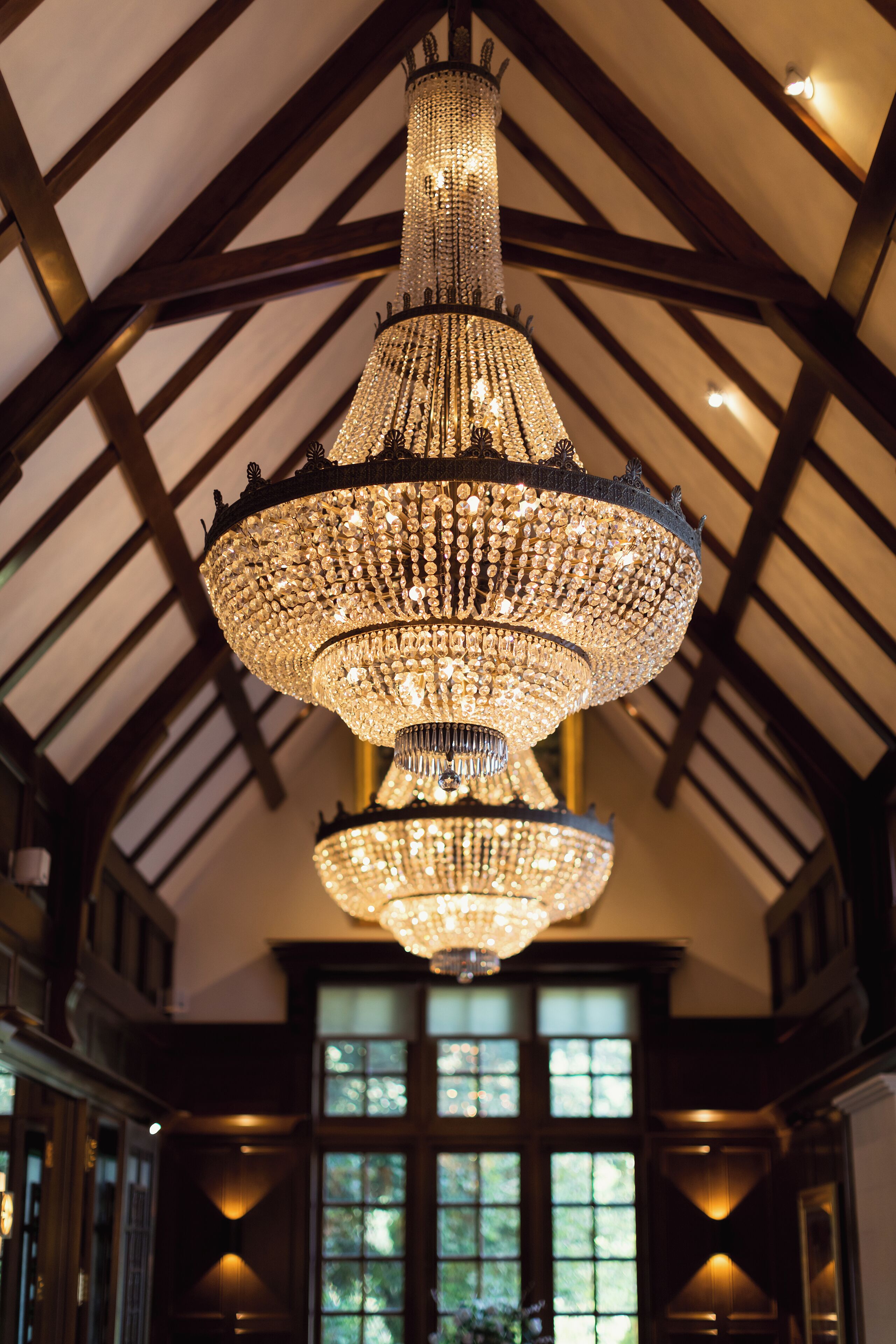 A grand chandelier with multiple tiers hanging from a high ceiling in a room with wooden beams and large windows.