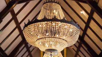 A grand chandelier with multiple tiers hanging from a high ceiling in a room with wooden beams and large windows.