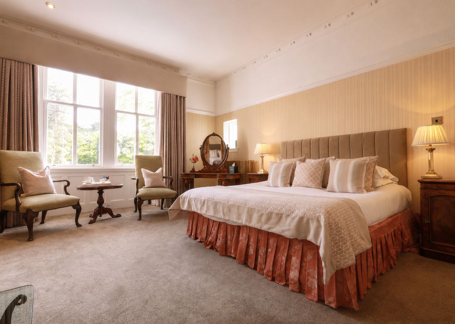 Habitación de hotel con una cama grande, dos sillones, una mesita, un aparador y una ventana con cortinas.